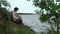 A teenager plays a guitar while sitting on the shore of a lake.