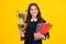 Teenager school girl with award winner trophy. Child hold books with gold trophy or winning cup isolated on yellow