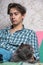 A teenager is sitting on the couch and stroking a gray cat sitting next to him