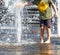 A teenager stands barefoot by a fountain and tries to stop a powerful stream of water with his hand on a bright hot day