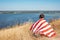 A teenager wrapped in an American flag sits in nature overlooking the bay. View from the back. Freedom concept