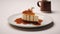 a tempting caramel Biscoff cheesecake displayed on a white table, accompanied by a slice and a cup of tea or coffee,the