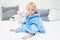 Ten month blond baby boy sitting on bed in blue bathrobe and looking at camera. White pillows in background. Baby care concept,