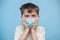 terrified boy in medical protective mask grabbed face with hands. hild with flu, influenza or cold protected from viruses, in bad