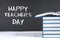 Text chalk on a chalkboard: Happy Teacher\'s Day. School supplies, office, books, apple.