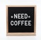 Text in english spelling `Need Coffee` with smiley face emojis on black felt board in a wooden frame. Letter Board on white