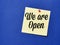 Text we are open written on yellow paper note with pin board over blue background.