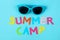 Text SUMMER CAMP of multicolored paper letters and sunglasses on a bright blue background. top view. flat lay