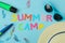 Text SUMMER CAMP of multicolored paper letters and sunglasses, hat and paperclip against a bright blue background. top view. flat