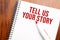 Text TELL US YOUR STORY written in notepad, Office wood table from above, concept image for blog title or header image. Aged