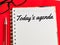 Text Today`s agenda on note book with pen and eye glasses  on red background.