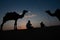 Thar desert, Rajasthan, India- 15.10.2019 : Silhouette of two cameleers and their camels at sand dunes. Cloud with setting sun,