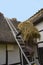 A thatcher with a bundle of straw on his back going up a ladder to a thatched roof on a half timber house to repair