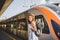 Theme railway and travel. Portrait young caucasian woman with toothy smile standing at train station train background with backpac