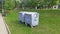 There are metal garbage containers on the grassy lawn near the tiled footpath. There are a bench and an urn nearby, trees grow, an
