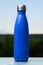 Thermo stainless bottle, sprayed with water. Sky and forest on background. On the glass desk. Thermos of matte blue color.
