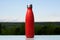 Thermo stainless bottle, sprayed with water. Sky and forest on background. On the glass desk. Thermos of matte red color.