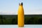 Thermo stainless bottle, sprayed with water. Sky and forest on background. On the glass desk. Thermos of matte yellow color.