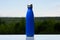 Thermo stainless bottle for water, tea or coffee. Sky and forest on background. On the glass desk. Thermos of matte blue color.