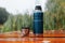 Thermos and mug with hot drink standing on wet wooden table after rain outdoors, close-up. Steam rises from thermo mug