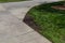 Thick grass and dark soil beside the turn in a concrete sidewalk, cutting corners visual metaphor, creative copy space, horizontal