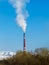 Thick white smoke comes out of the red pipe of the factory against a clear blue sky, snowy mountains and treetops. Vertical orient