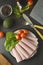 Thin slices of ham rolled on plate with fresh vegetables, dark background. Breakfast food, ingredient for sandwich. Flat lay food