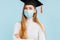 Thinking girl graduate in a medical mask, student in a graduation hat thinks and looks to the side on a blue background