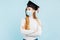 Thinking girl graduate in a medical mask, student in a graduation hat thinks and looks to the side on a blue background