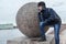 Thinking teenage boy dressed warm jacket and hat standing near big stone ball. Embankment of Neva river on the spit of Vasilyevsky