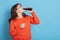 Thirsty female wearing orange jumper standing isolated over blue background  holding bottle of fizzy water and drinking  adorable