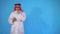 Thoughtful arab man standing on a blue background uses the phone