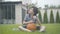 Thoughtful Caucasian boy sitting on green grass with ball as blurred brother climbing on basketball hoop at the