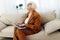 a thoughtful, enthusiastic elderly woman in a stylish brown suit sits on a beige sofa with a laptop on her lap and looks