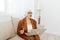 a thoughtful, enthusiastic elderly woman in a stylish brown suit sits on a beige sofa with a laptop on her lap and looks