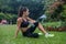 Thoughtful fitness girl sitting on grass in park resting after exercising or running, listening to music and holding a