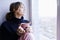 A thoughtful girl in a blue sweater sits by the window with a Cup of coffee