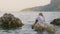 Thoughtful girl teenager in white wet tunic sitting on stone in sea on mountain landscape. Tourist girl sitting on rock