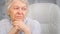 Thoughtful grey haired senior lady looks into window holding head over hands and sitting on white chair in office