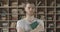 Thoughtful handsome young man standing in library with book in hands and looking at camera. Portrait of serious