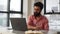 Thoughtful Indian businessman in glasses using laptop writing notes in planner notebook sitting at office table on