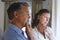 Thoughtful senior caucasian couple standing next to window, holding their chins