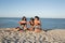 Three beautiful girls in jean shorts and black bras are sitting on the sand near the sea and holding watermelon slices.