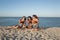 Three beautiful girls in jean shorts and black bras are sitting on the sand near the sea and holding watermelon slices.