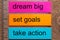 Three bookmark stickers with the words dream big, set goals, take action on a dark natural wooden table