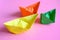 Three bright colorful multicolor paper origami boats in orange, yellow and green colors on a pink background..