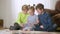 Three charming happy Caucasian children using tablet talking and smiling sitting in living room. Positive boys and girl