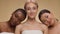 Three cheerful multiethnic female friends wrapped in towels and smiling at camera together, posing over beige background