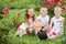 Three children sit on grass among bushes of roses