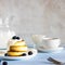 Three cottage cheese pancakes one on another topped with cream and blueberries on light background and defocused cup of coffee,